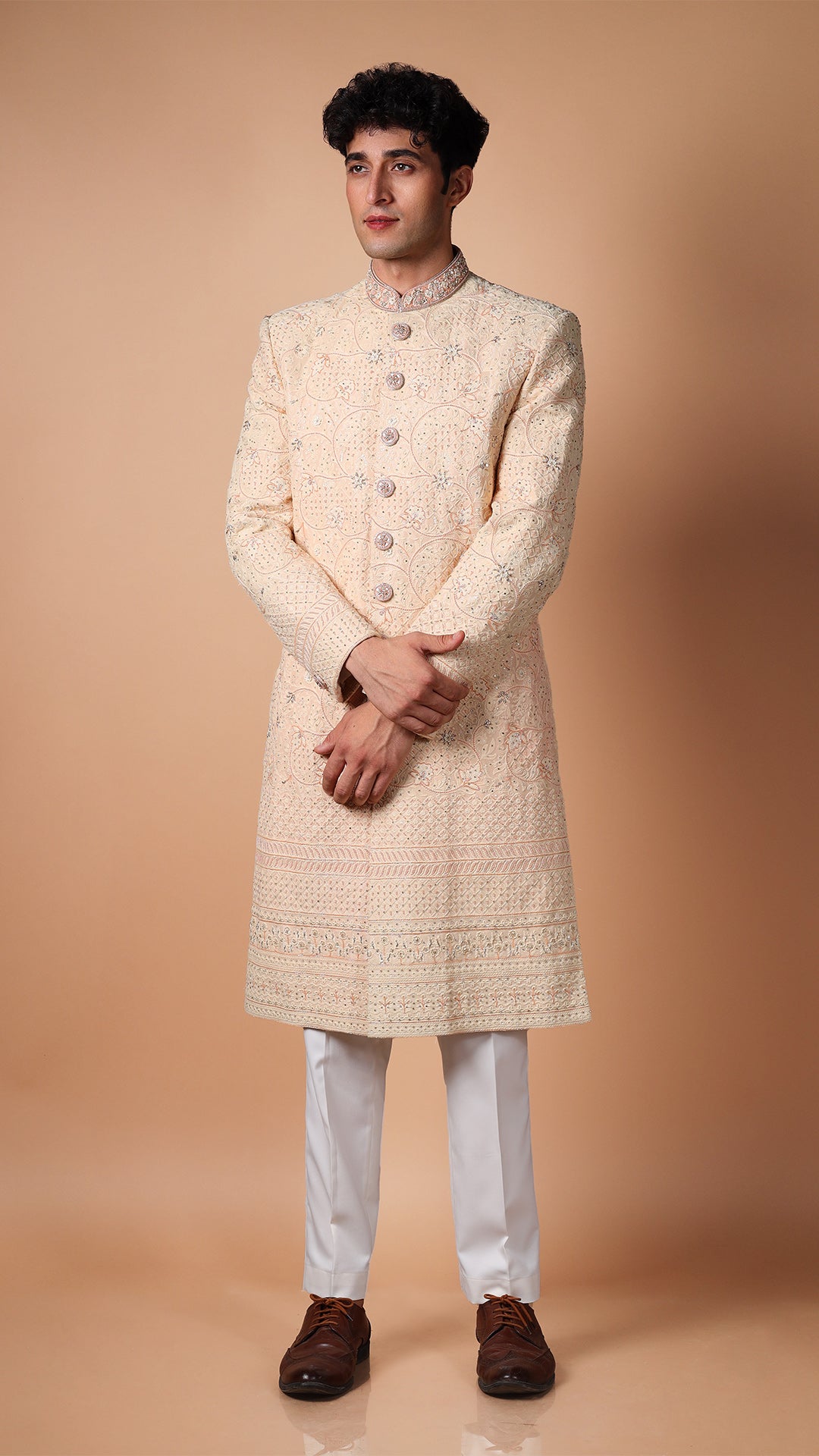 The model stands confidently in the Peach Lucknowi Hand Embroidered Sherwani, paired with off-white bottoms. The intricate Lucknowi embroidery on the luxurious fabric highlights traditional craftsmanship.